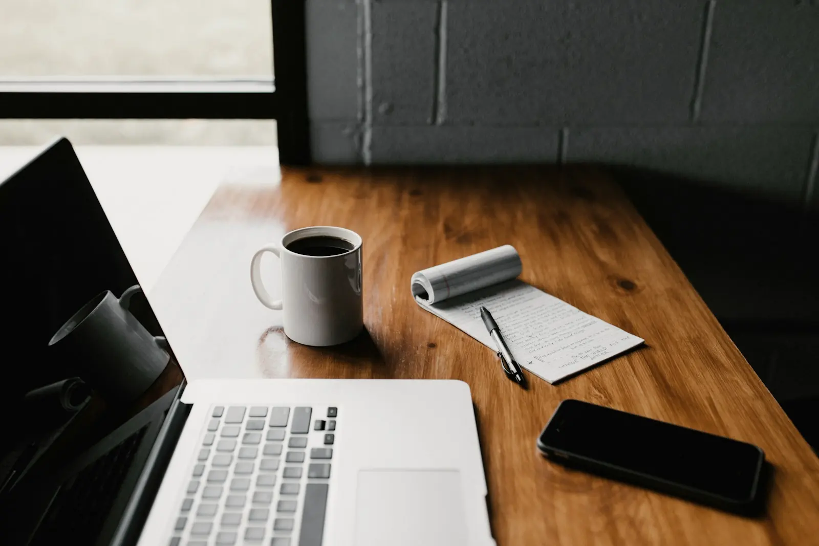 A laptop, notebook, coffee and phone on a dark wooden desk