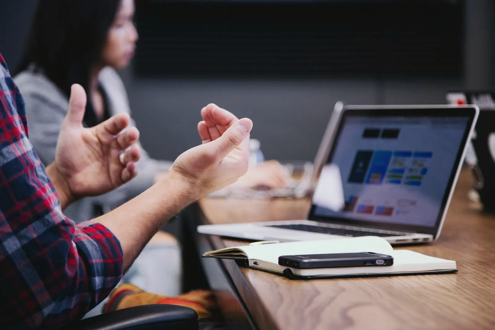 A person explaining a point with open hands across a table, a laptop and notebook in front of them